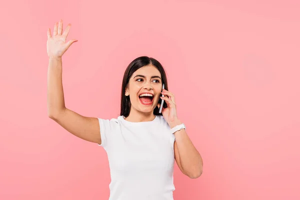 Excitada chica morena bonita hablando en el teléfono inteligente agitando la mano aislado en rosa - foto de stock