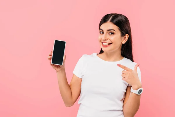 Sonrisa bonita chica morena apuntando con el dedo en el teléfono inteligente con pantalla en blanco aislado en rosa - foto de stock