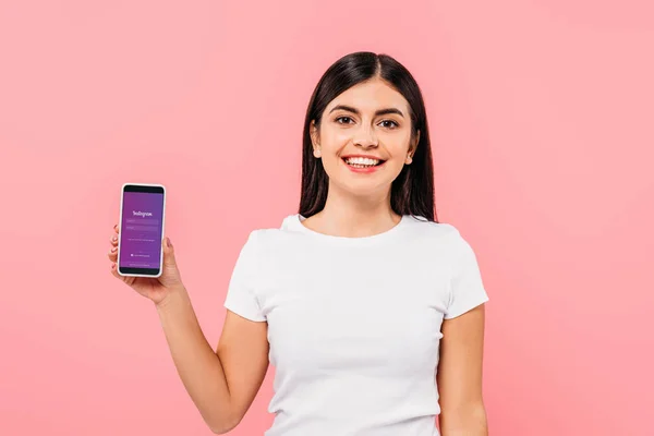 KYIV, UCRANIA - 20 de septiembre de 2019: una bonita chica morena sonriente sosteniendo el teléfono inteligente con la aplicación Instagram aislada en rosa - foto de stock