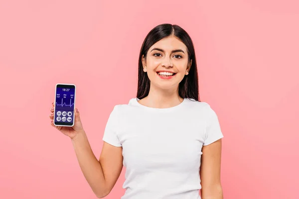 Sonriente chica morena bonita sosteniendo teléfono inteligente con aplicación sanitaria aislado en rosa - foto de stock
