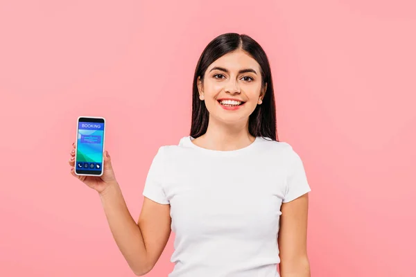 Sonriente chica morena bonita sosteniendo teléfono inteligente con aplicación de reserva aislado en rosa - foto de stock
