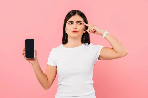 Nachdenkliche hübsche brünette Mädchen hält Smartphone mit leerem Bildschirm isoliert auf rosa — Stockfoto