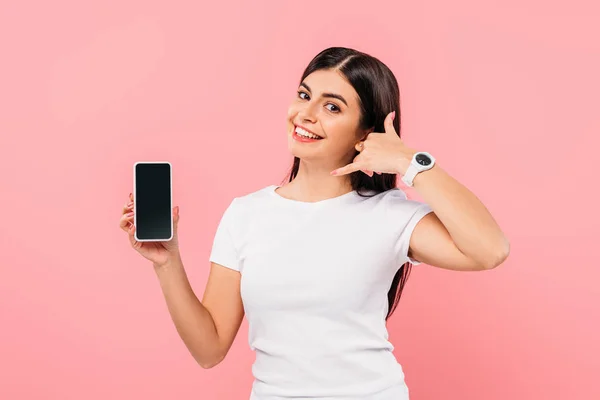 Sonriente chica morena bonita sosteniendo teléfono inteligente con pantalla en blanco y mostrando gesto de llamada aislado en rosa - foto de stock