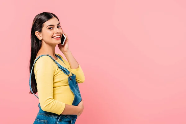 Seitenansicht eines lächelnden schwangeren hübschen Mädchens, das auf dem Smartphone mit der Hand auf dem Bauch spricht, isoliert auf rosa — Stockfoto