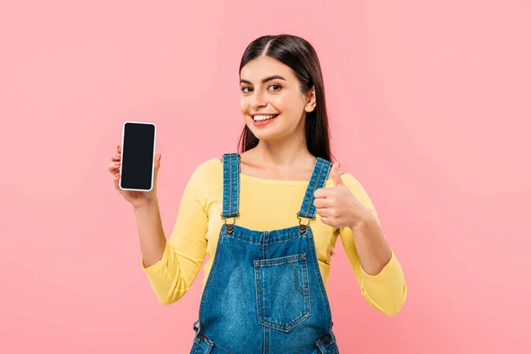 Усміхнена красива дівчина тримає смартфон з порожнім екраном і показує великий палець вгору ізольовано на рожевому — стокове фото