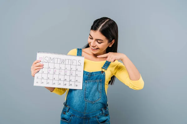 Sorrindo grávida bonita menina apontando com o dedo no calendário período isolado em cinza — Fotografia de Stock