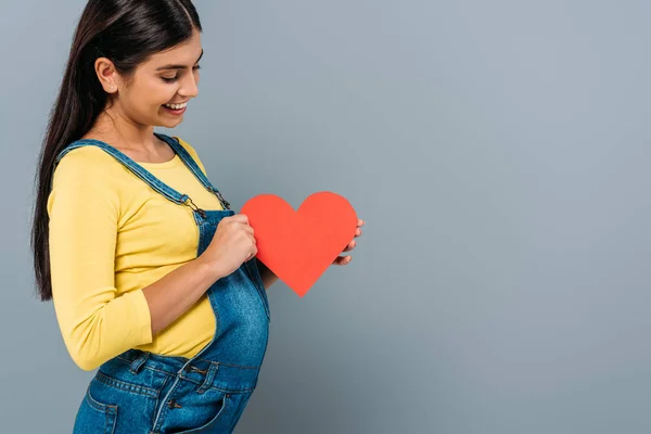 Vista laterale di sorridere incinta bella ragazza in possesso di cuore di carta isolato su grigio — Foto stock