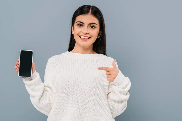 Sonriente chica morena bonita en suéter blanco apuntando con el dedo al teléfono inteligente con pantalla en blanco aislado en gris - foto de stock