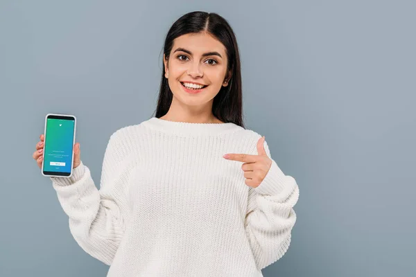 KYIV, UCRANIA - 20 de septiembre de 2019: una guapa morena sonriente en suéter blanco apuntando con el dedo al teléfono inteligente con la aplicación de twitter aislada en gris - foto de stock