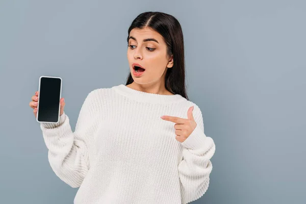 Shocked pretty brunette girl in white sweater pointing with finger at smartphone with blank screen isolated on grey — Stock Photo