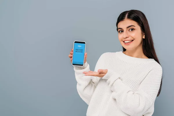 KYIV, UCRANIA - 20 DE SEPTIEMBRE DE 2019: bonita chica morena sonriente en suéter blanco sosteniendo smartphone con aplicación skype aislada en gris - foto de stock