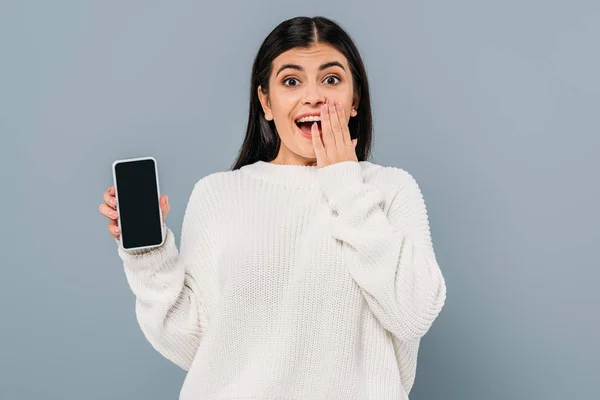 Excitada chica morena bonita en suéter blanco sosteniendo teléfono inteligente con pantalla en blanco aislado en gris - foto de stock