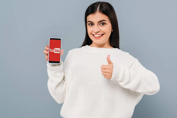 Lächelndes hübsches Mädchen im weißen Pullover zeigt Smartphone mit Youtube-App und Daumen hoch vereinzelt auf grau — Stockfoto