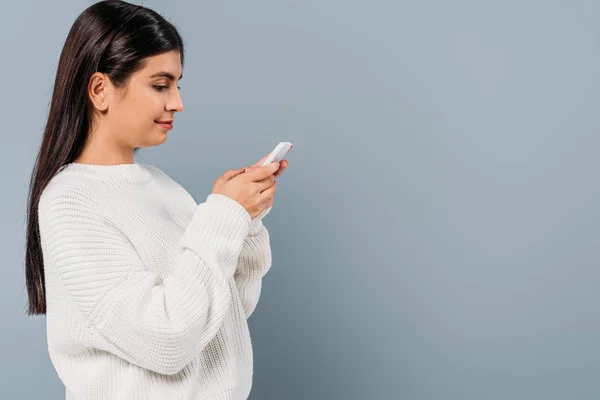 Side view of smiling pretty brunette girl in white sweater using smartphone isolated on grey — Stock Photo