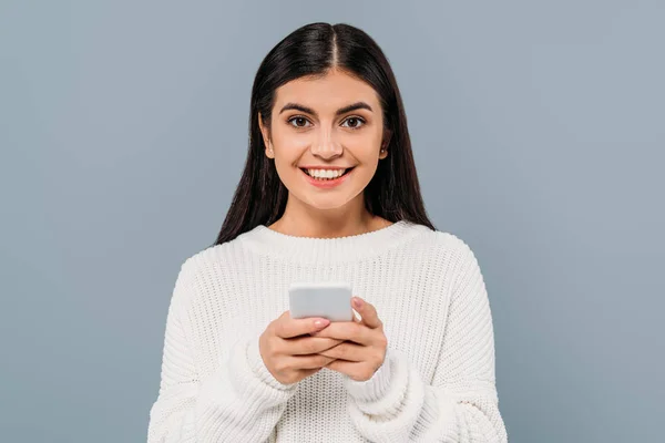 Sonrisa bonita chica morena en suéter blanco usando teléfono inteligente aislado en gris - foto de stock