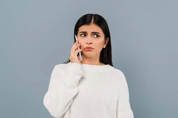 Trauriges hübsches brünettes Mädchen im weißen Pullover, das auf dem Smartphone isoliert auf grau spricht — Stockfoto