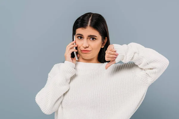 Triste bella ragazza bruna in maglione bianco parlando su smartphone e mostrando pollice verso il basso isolato sul grigio — Foto stock