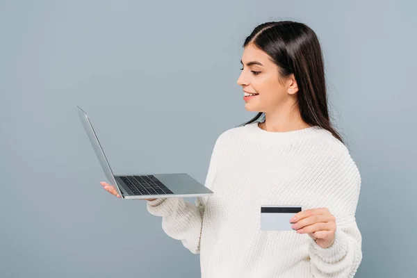 Sonriente chica bonita en suéter blanco sosteniendo portátil y tarjeta de crédito aislado en gris - foto de stock