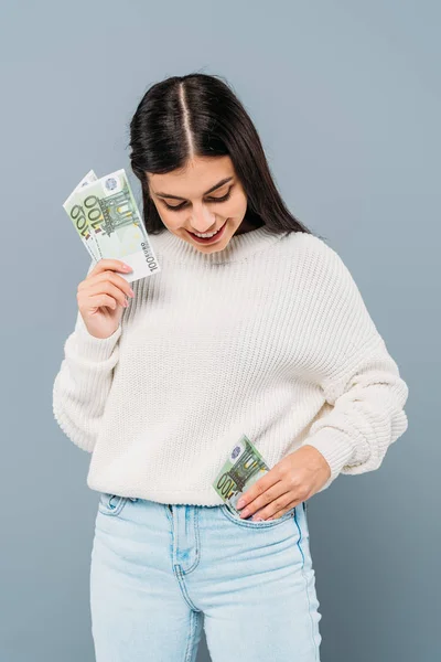 Усміхнена красива дівчина в білому светрі кладе банкноти євро в кишеню ізольовані на сірому — стокове фото