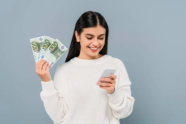 Усміхнена красива дівчина в білому светрі тримає банкноти євро і використовує смартфон ізольовано на сірому — стокове фото