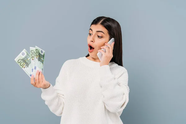 Impactado chica bonita en suéter blanco con billetes en euros y hablando en el teléfono inteligente aislado en gris - foto de stock