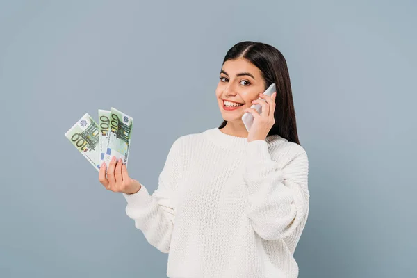 Chica bonita sonriente en suéter blanco sosteniendo billetes en euros y hablando en un teléfono inteligente aislado en gris - foto de stock