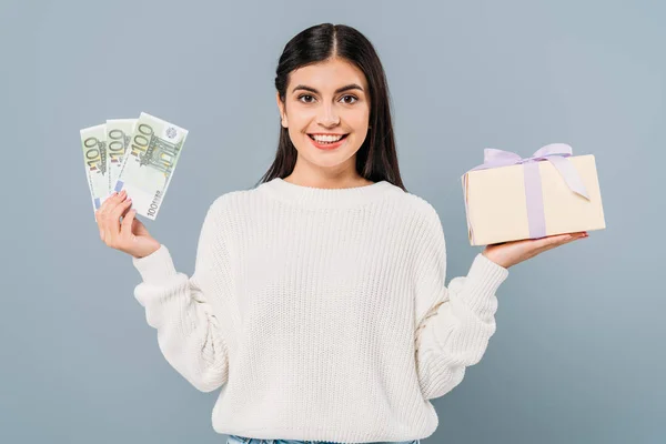 Jolie fille souriante en pull blanc tenant billets en euros et boîte cadeau isolé sur gris — Photo de stock