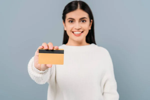 Избирательный фокус улыбающейся красивой девушки в белом свитере, представляющей кредитную карту, изолированную на серой — стоковое фото