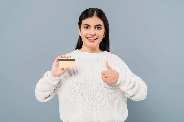 Smiling pretty girl in white sweater holding credit card and showing thumb up isolated on grey — Stock Photo