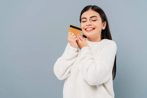 Sonriente chica bonita en suéter blanco celebración de la tarjeta de crédito con los ojos cerrados aislados en gris - foto de stock