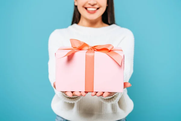 Recortado vista de sonriente chica bonita en suéter blanco presentando caja de regalo aislado en azul - foto de stock
