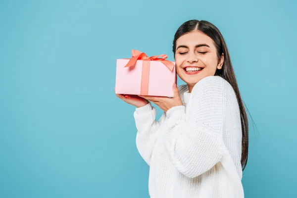 Sonriente chica bonita en suéter blanco celebración caja de regalo con los ojos cerrados aislados en azul - foto de stock