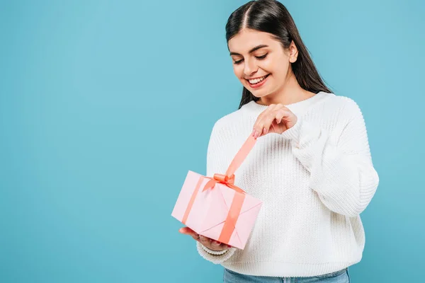 Smiling pretty girl in white sweater unwrapping gift box isolated on blue — Stock Photo