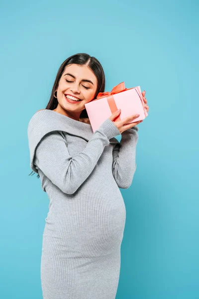 Улыбающаяся беременная красивая девушка держит подарок с закрытыми глазами изолированы на синий — стоковое фото