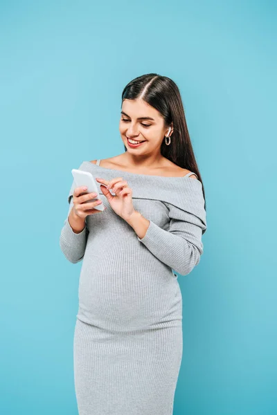 Chica bonita embarazada sonriente usando teléfono inteligente aislado en azul - foto de stock