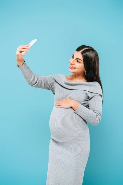 Sorridente incinta bella ragazza prendendo selfie su smartphone isolato su blu — Foto stock