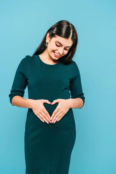 Усміхнена вагітна дівчина показує серцевий жест на животі ізольовано на синьому — стокове фото