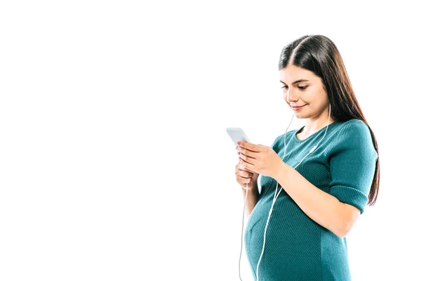 Niña embarazada sonriente usando smartphone y escuchando música en auriculares aislados en blanco - foto de stock