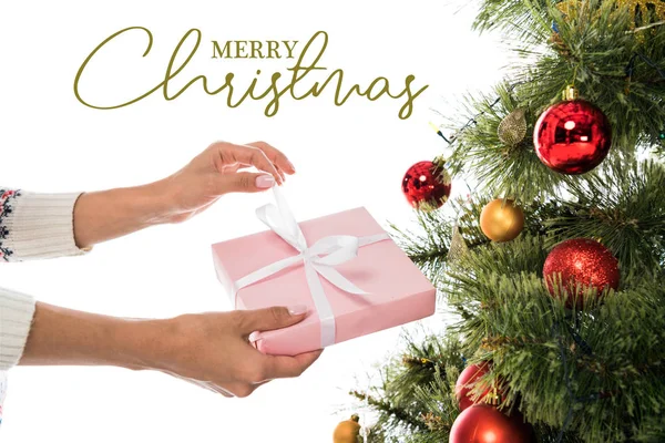 Cropped view of woman holding pink present near christmas tree isolated on white with merry Christmas lettering — Stock Photo