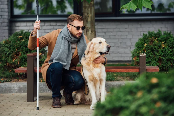 Слепой мужчина с тростью обнимает собаку-поводыря на улице — стоковое фото