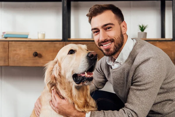 Smiling man looking at camera while petting golden retriever at home — Stock Photo