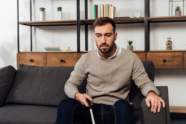 Hombre con discapacidad visual sosteniendo bastón mientras está sentado en el sofá en la sala de estar - foto de stock