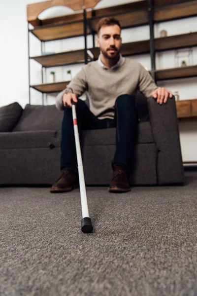 Enfoque selectivo del hombre con discapacidad visual que sostiene el bastón mientras está sentado en el sofá en la sala de estar - foto de stock