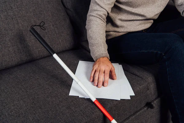 Cropped view of blind man reading braille font beside walking stick on sofa — Stock Photo