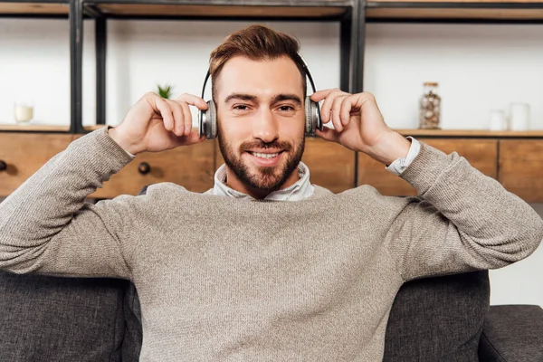 Uomo sorridente in cuffia seduto sul divano in soggiorno — Foto stock