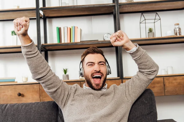 Exited man in headphones with hands in air on sofa in living room — Stock Photo