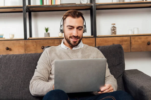 Homme souriant dans les écouteurs en utilisant un ordinateur portable sur le canapé dans le salon — Photo de stock