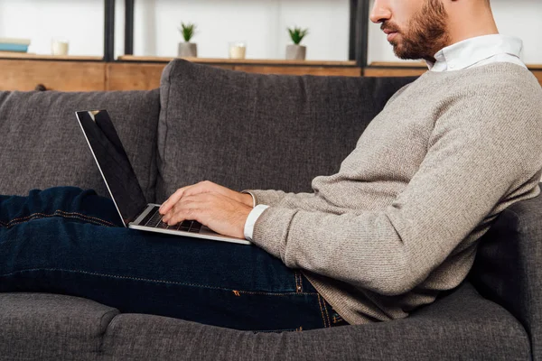 Vista recortada del hombre escribiendo en el teclado del ordenador portátil en el sofá en casa - foto de stock