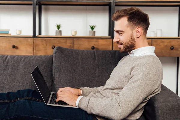 Hombre sonriente escribiendo en el teclado del ordenador portátil en el sofá en la sala de estar - foto de stock