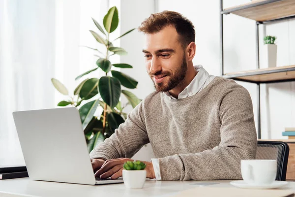 Homme souriant utilisant un ordinateur portable à côté d'une tasse de café sur la table — Photo de stock